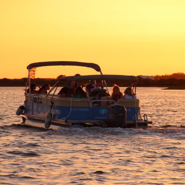 A pontoon - catamaran boat with happy guests cruising on the Ria Formosa Natural Park at sunset, with the sky and water glowing in warm shades of orange.