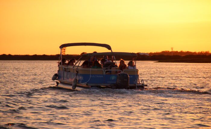 A pontoon - catamaran boat with happy guests cruising on the Ria Formosa Natural Park at sunset, with the sky and water glowing in warm shades of orange.