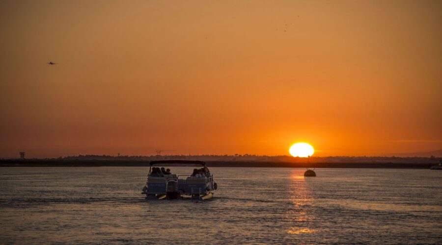 A boat with passengers cruising on the Formosa Natural Park at sunset, with the sun low on the horizon casting an orange glow across the sky and water.
