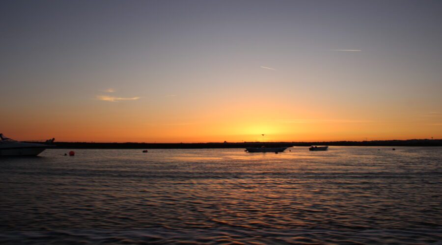 Boats floating on the Ria Formosa Natural Park during sunset, with the sky displaying shades of orange and yellow, and the water reflecting the warm colors.